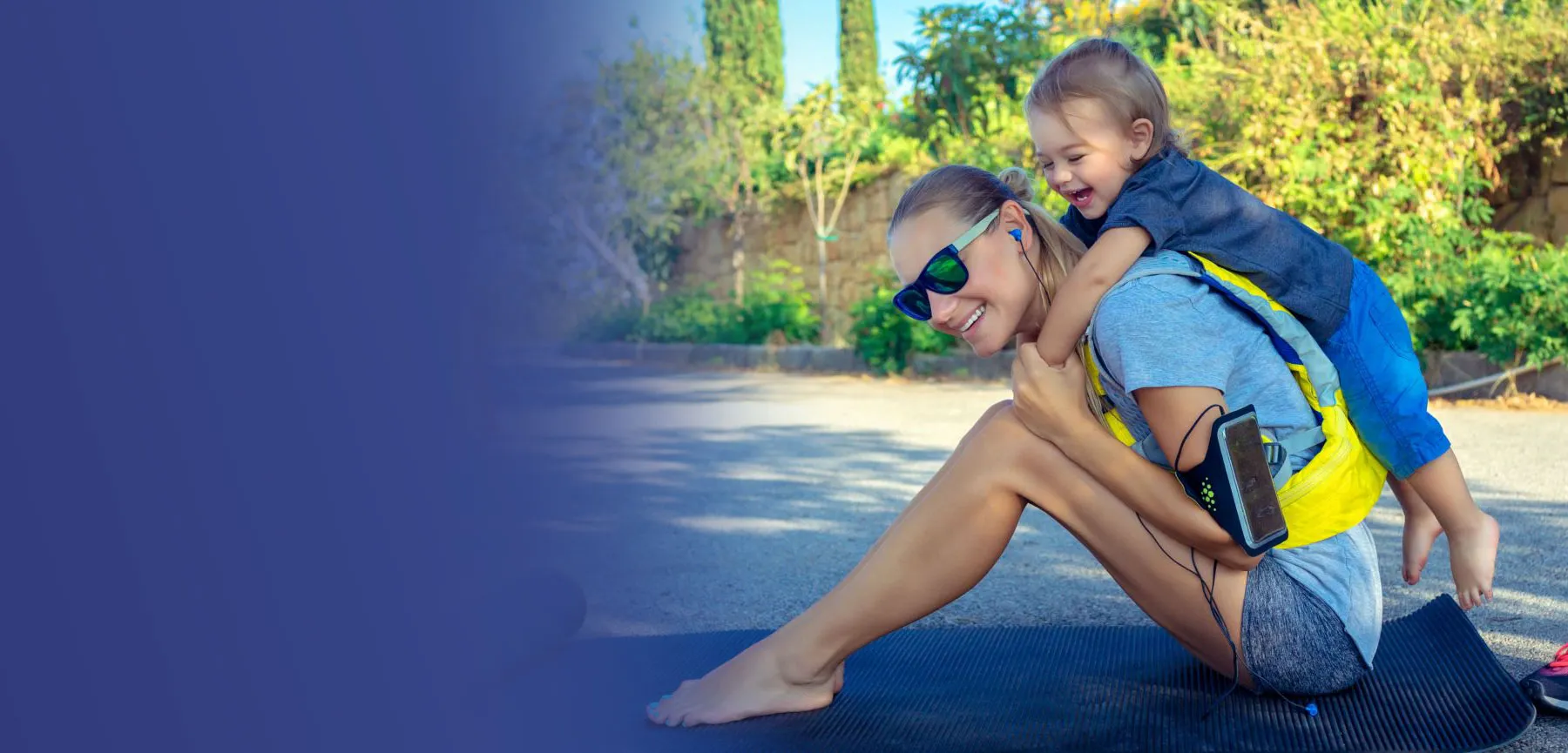 A mother wearing sunglasses gives her laughing toddler a piggyback ride outdoors